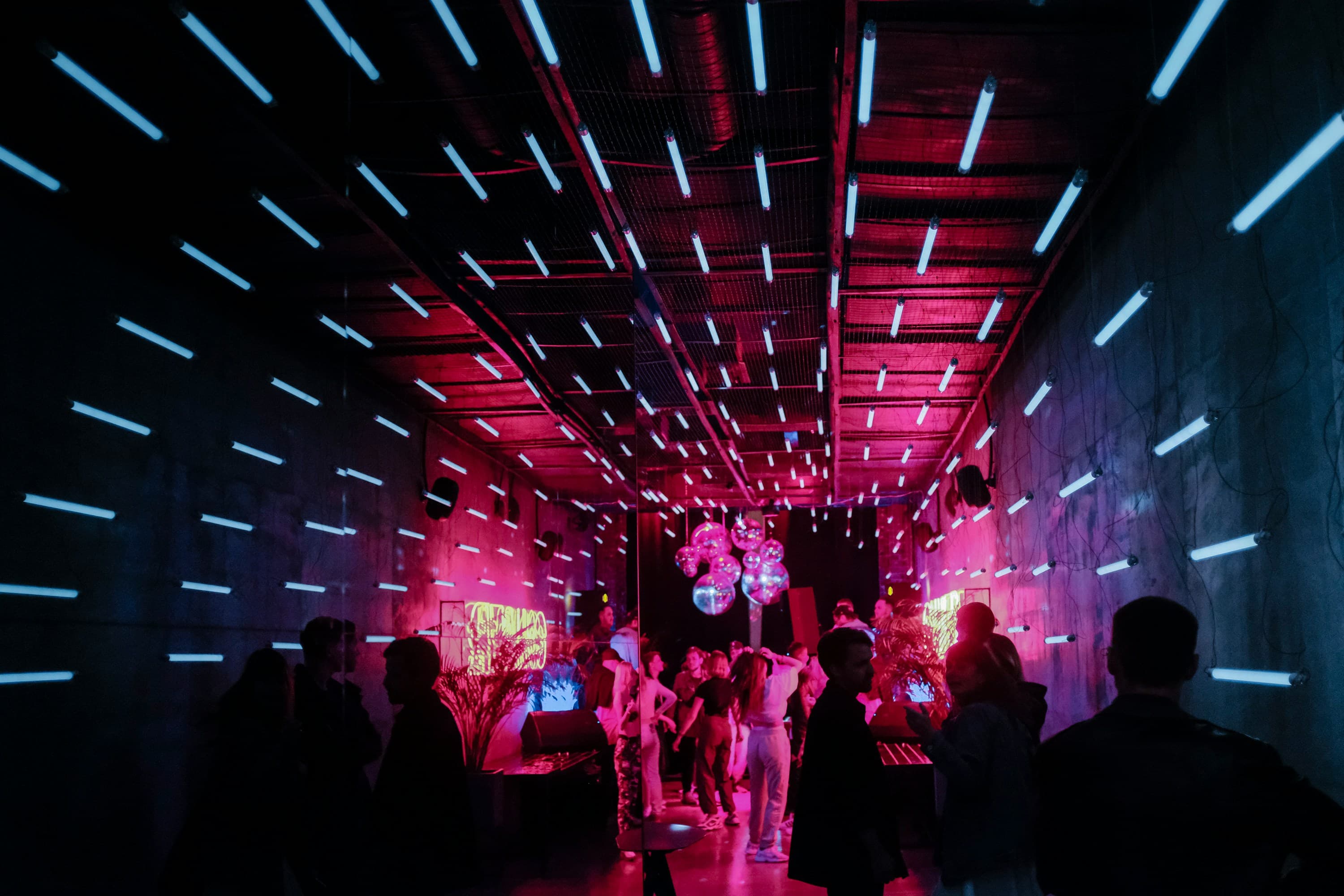 Menschen feiern bei einem Event mit Neonlichtern an den Wänden und der Decke, während bunte Neonschilder, Discokugeln und rosa-blaues Licht die Szene erhellen